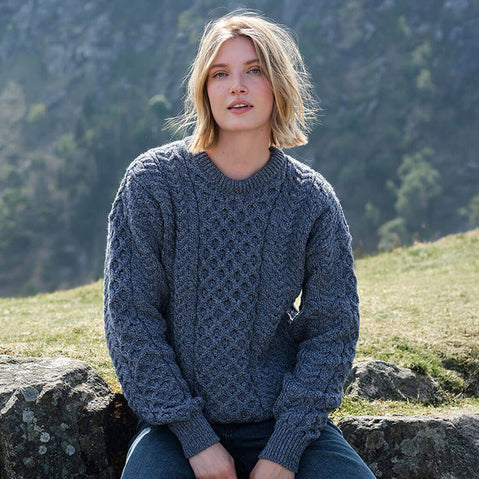 Woman wearing a denim blue Aran sweater sitting on a rock with a mountainous background