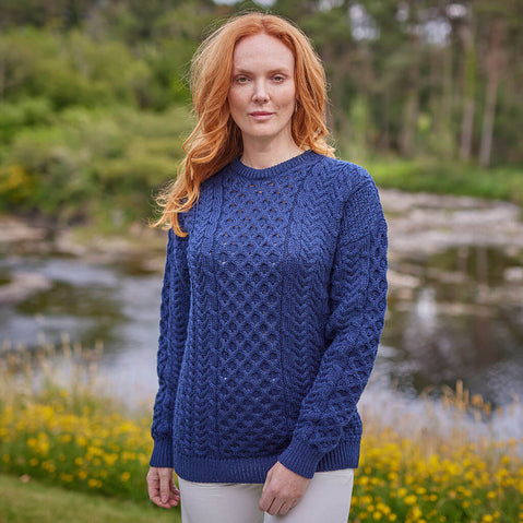 Woman wearing a blue knitted sweater standing in a natural setting with a stream and greenery.