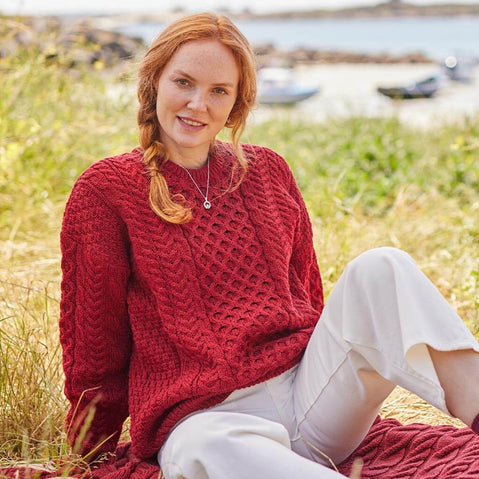 A red- headed woman wearing a red Aran sweater sitting on grass by a body of water.