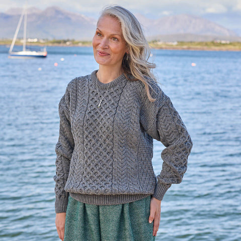 Woman wearing a gray cable knit sweater by a body of water with mountains in the background