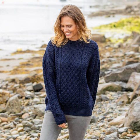 A woman standing by a body of water wearing a traditional Aran sweater with a crew neck design.