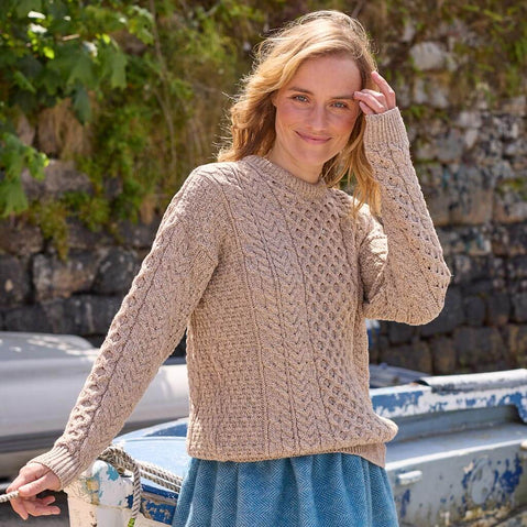 Woman wearing a beige knitted sweater standing outdoors with boats and stone wall in the background