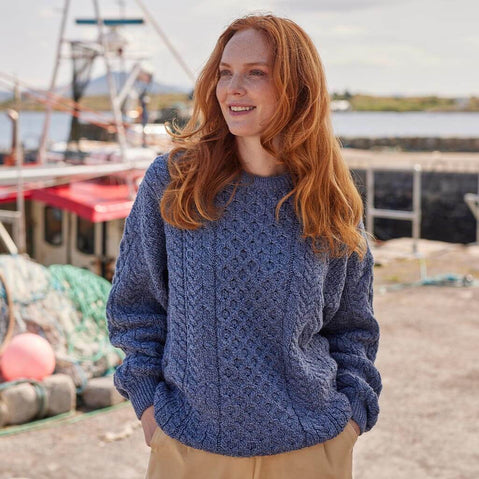 A red headed woman wearing a blue Aran sweater standing by a harbor with boats in the background