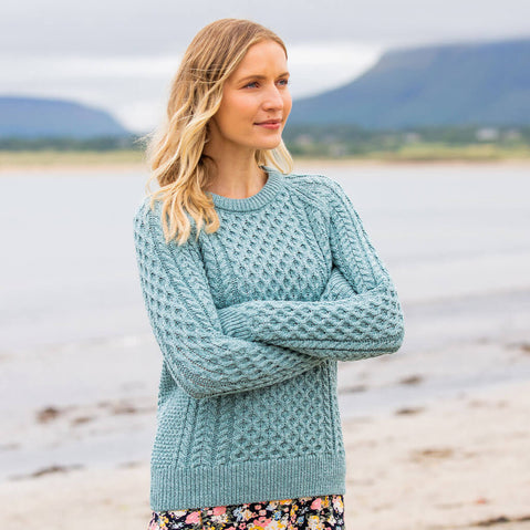Woman wearing a teal knitted sweater standing on a beach with mountains in the background