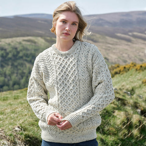 Woman wearing an oatmeal colored Aran sweater standing in a scenic outdoor setting with hills and greenery.