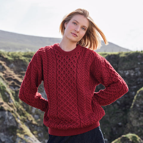 Image featuring the Women's Traditional Crew Neck Aran Sweater in red, crafted with authentic Irish Aran cable knit patterns and a timeless crew neck silhouette, showcasing traditional Irish craftsmanship in a rugged coastal setting.
