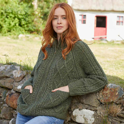 Woman wearing a green Women's Hand Knit Aran Zipper Cardigan sitting on a stone wall outdoors.