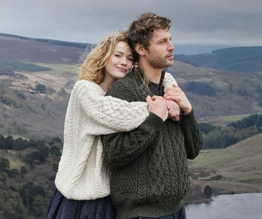 A man and woman stand together outdoors, wrapped in warm cable-knit aran sweaters. The woman hugs the man from behind and smiles, while he looks off into the distance. The background features rolling hills under a cloudy sky.