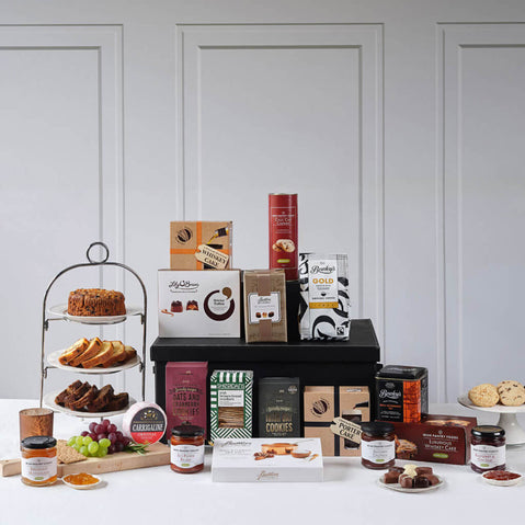 Finest Irish Foods Gift Basket. An assortment of packaged gourmet foods arranged on a table. In the foreground, jars of spreads and condiments sit beside various baked goods, including cookies and pastries. A multi-tiered stand displays more treats, while boxes of different snacks are stacked in the background. The setting features a simple white backdrop for emphasis on the products.
