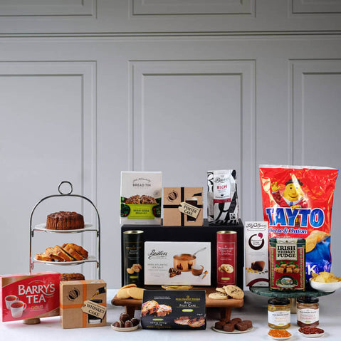 Favorite Irish Classics Gift Basket. A festive display featuring an assortment of food items arranged on a table. In the foreground, there's a multi-tiered stand with baked goods and treats. Several boxes of tea and snacks are visible, including Barry's Tea and Tayto crisps.
