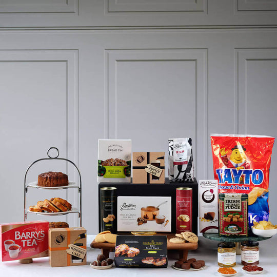 Traditional Irish Feast Hamper. A festive display featuring an assortment of food items arranged on a table. In the foreground, there's a multi-tiered stand with baked goods and treats. Several boxes of tea and snacks are visible, including Barry's Tea and Tayto crisps.