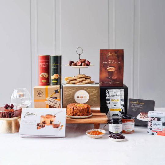 Best of Ireland Gourmet Gift Basket. A display of assorted gourmet Irish treats, including a tiered stand with cookies, various packages of chocolates, jars of preserves, and a round cake. There are also recipe cards and a wine glass, set against a light background.