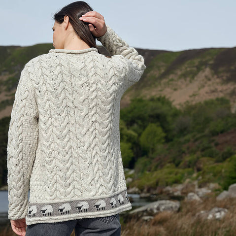 Back view of a person wearing an oatmeal-colored Women's Sheep Trim Aran Tunic Wool Sweater with a cable-knit design and a row of small sheep patterns along the bottom hem, standing outdoors with hills and greenery in the background.