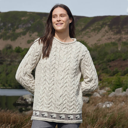 A person wearing an oatmeal-colored Women's Sheep Trim Aran Tunic Wool Sweater with a cable-knit design and a row of small sheep patterns along the bottom hem, standing outdoors with hills and greenery in the background