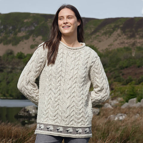 A person wearing an oatmeal-colored Women's Sheep Trim Aran Tunic Wool Sweater with a cable-knit design and a row of small sheep patterns along the bottom hem, standing outdoors with hills and greenery in the background