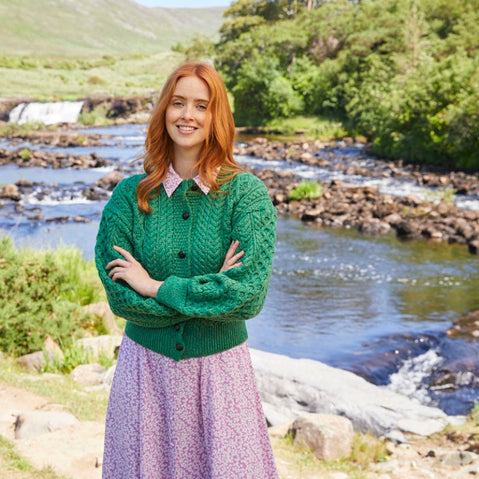 A woman stands by a river, smiling, arms folded and wearing a green knitted Aran cardigan with buttons. She pairs it with a pink floral skirt, and the backdrop features greenery