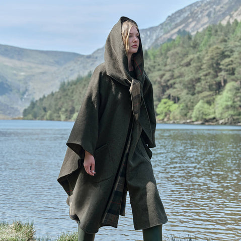 Person wearing a hooded Irish walking cape by a lake with mountains in the background