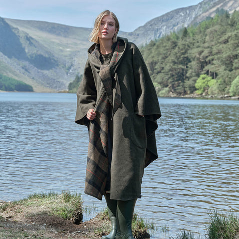 Woman in a green Irish walking cape standing by a lake with mountains in the background