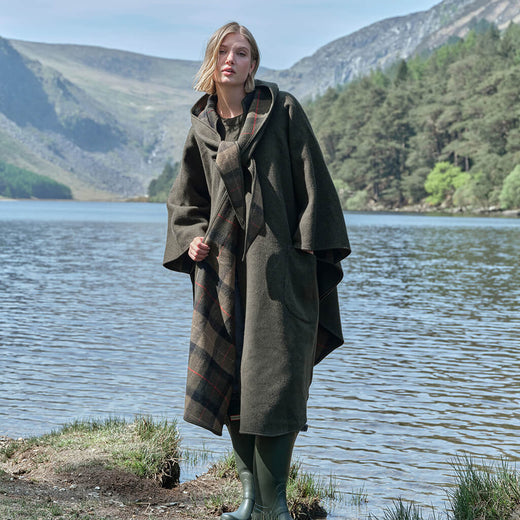 Woman in a green Irish walking cape standing by a lake with mountains in the background