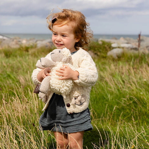A young child with curly hair stands in a grassy field, smiling while holding a stuffed sheep toy. The background features a coastline and cloudy sky. The child is dressed in a cozy kids Aran cardigan over a blue dress.
