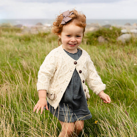 A young child with curly hair wears a light-colored knitted kids Aran cardigan and a blue dress. She is playing in a grassy field, smiling and enjoying the outdoors, with a coastline visible in the background.