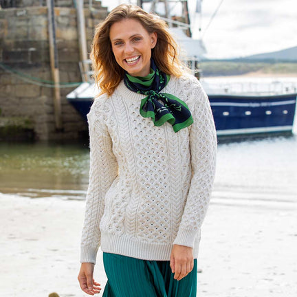Woman wearing a cream Women's Crew Neck Irish Wool Classic Sweater and green scarf by a body of water with a boat in the background