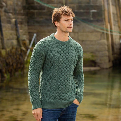 Man wearing a green Aran sweater standing by a body of water with stone walls in the background.