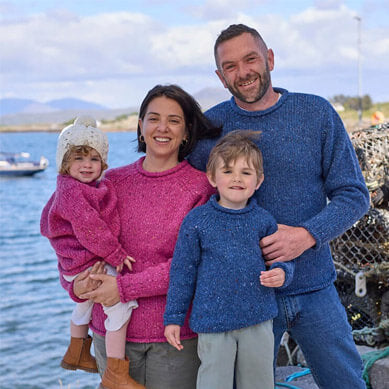 A family of four wearing matching pink and blue speckled woolen sweaters, smiling together at a coastal harbor with mountains and a boat in the background.