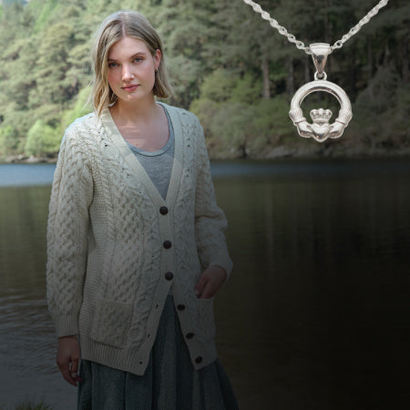Woman wearing a patterned cardigan with a necklace featuring a circular pendant with a crown design, standing by a body of water with trees in the background.