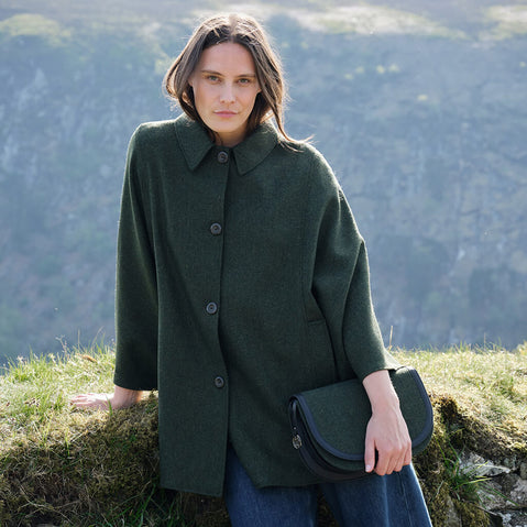 Woman in a green cape and matching handbag, standing on a grassy hill with mountains in the background