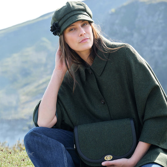 Woman in a green cape, matching hat and handbag, sitting outdoors with mountains in the background