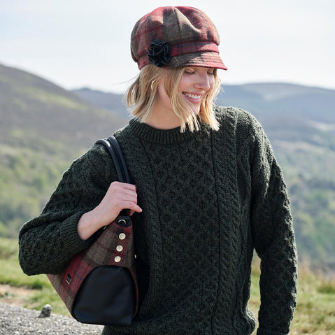 Woman wearing a dark green Aran sweater and plaid hat with a black bag, standing in a scenic outdoor setting.