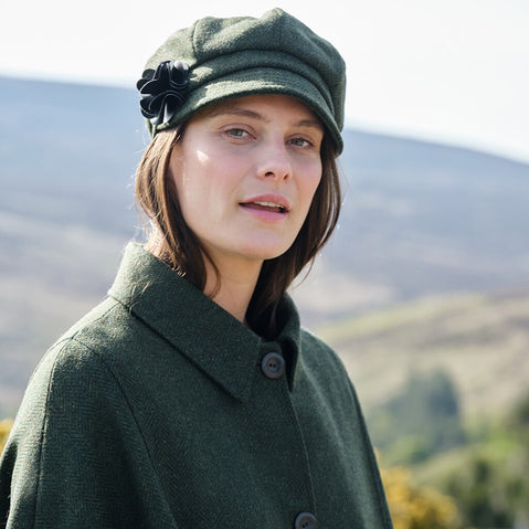 Woman wearing a green coat and hat with a flower, standing outdoors with a blurred natural background