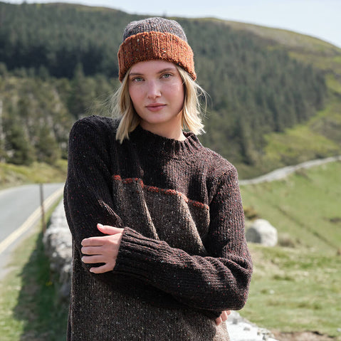 Woman wearing a brown knitted sweater and hat standing in a scenic outdoor setting with green hills and a road.