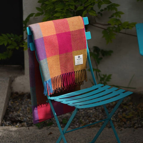 A colorful plaid blanket drapes over a turquoise metal chair, set against a backdrop of green foliage and a textured surface.
