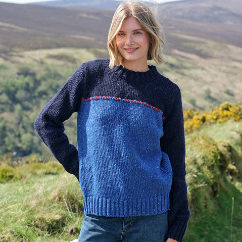 Woman wearing a blue and navy sweater standing in a scenic outdoor setting with hills and flowers.