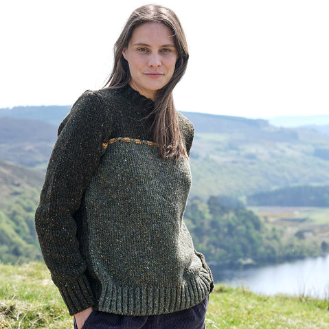 Woman wearing a green knitted sweater standing in a scenic outdoor setting with mountains and a lake.