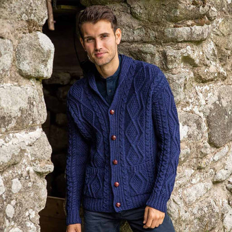 Man wearing a navy The Irish Store Men's Classic Aran Cardigan standing against a stone wall.