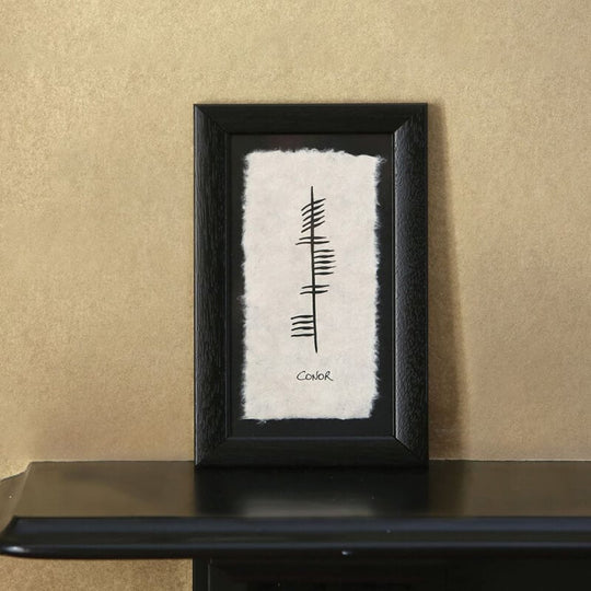 Ogham Personalized Name Plaque. A framed artwork displayed on a black surface, featuring a minimalist design of vertical lines and marks on textured paper. The word