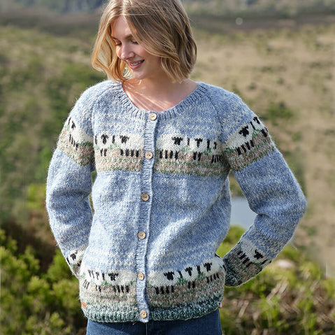 A person wearing a light blue Women's Mohair Sheep Cardigan with black, white, and green sheep and geometric patterns across the chest and sleeves. The cardigan features wooden buttons and a soft, fuzzy mohair texture, set against an outdoor backdrop of greenery and hills.