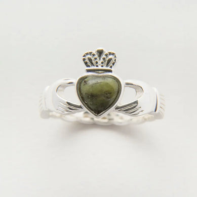 A silver Claddagh ring featuring a heart-shaped green Connemara Marble stone at its center, topped with a small crown. The band has intricate detailing on the sides, showcasing an elegant design. The background is plain and neutral, emphasizing the ring's features.