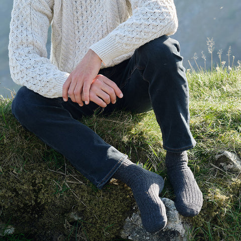 Person wearing a white Aran sweater, dark jeans and grey wool socks, while sitting on a grassy rock.