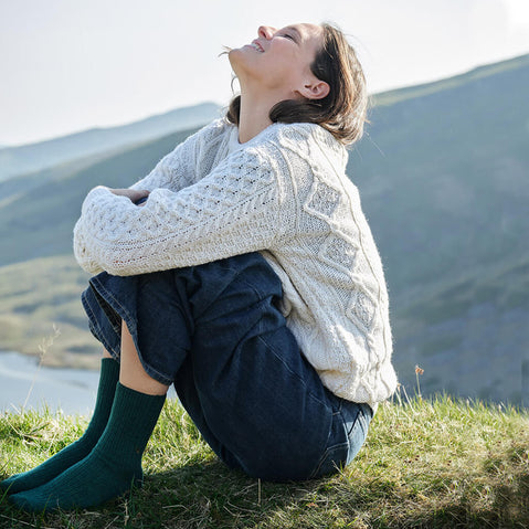 Woman sitting on a grassy hill wearing a white Aran sweater and blue jeans, green wool socks, and with mountains in the background.