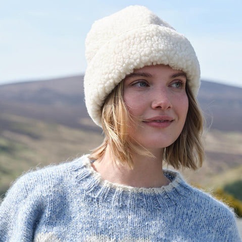 Woman wearing a white sheepswool hat and blue sweater outdoors with mountains in the background