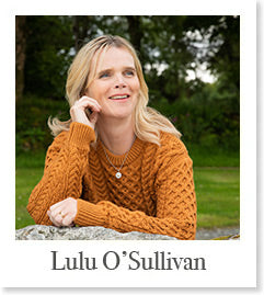 A woman with blonde hair sits on a stone wall, wearing an orange knitted sweater. She is smiling and looking off to the side, with greenery in the background. Below the image is the name