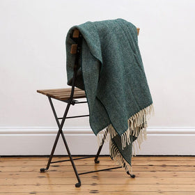 A green herringbone patterned Irish throw blanket draped over the back of a chair in a room with a plain wall and wooden flooring.