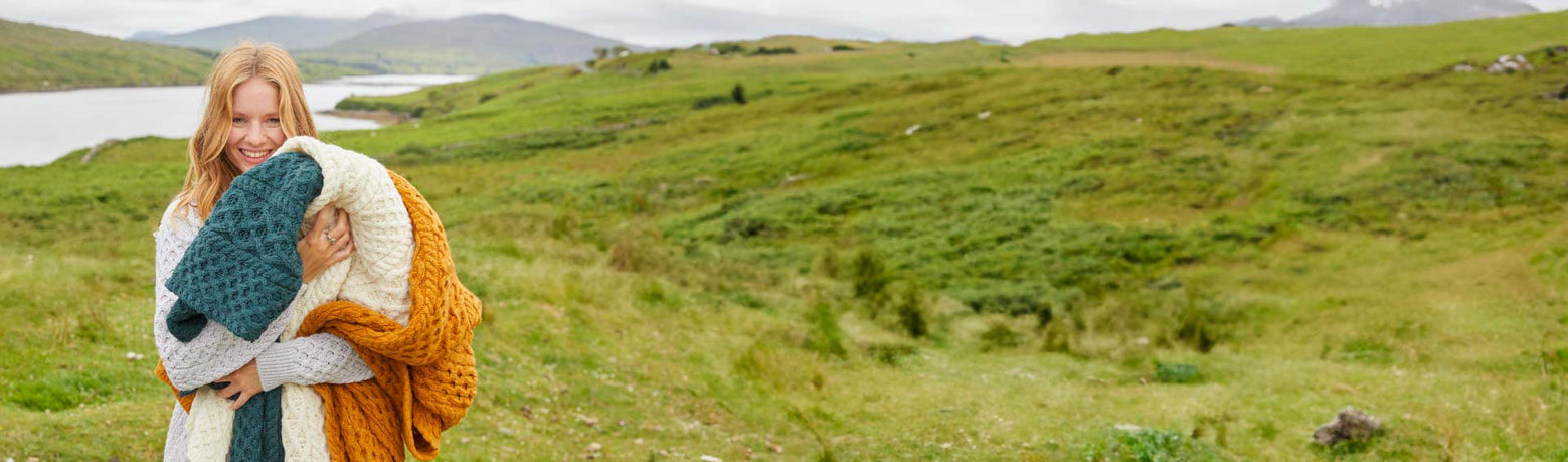 Woman holding a colorful knitted blanket with a scenic background of a lake and mountains.