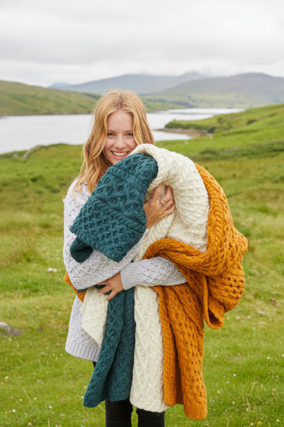 Woman holding a colorful knitted blanket with a scenic background of a lake and mountains.