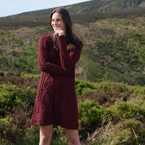 Woman in a burgundy Aran knit dress standing in a natural landscape with greenery and mountains.