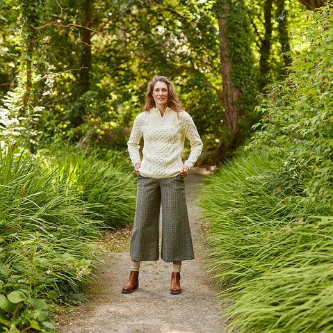 A woman stands on a path surrounded by lush greenery. She is wearing a light-colored Aran sweater and wide-legged gray trousers, with brown shoes. The background features dense foliage and trees, creating a serene atmosphere.